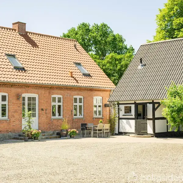 En murstensbygning med rødt tag står ved siden af en bindingsværksbygning. Gruset gårdsplads omgivet af grønne buske og træer. Der ses udendørs bord og stole tæt ved huset.