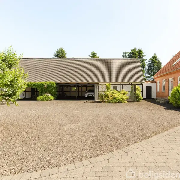 En gårdsplads med grus omgivet af en rød murstensbygning til højre og en garagebygning med grønne planter til venstre. En hvid bil er parkeret foran garagen.