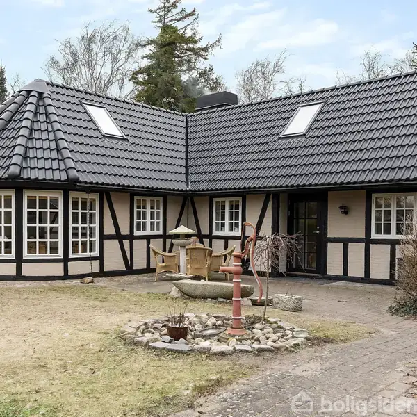 Et bindingsværkshus med sort tegltag står i en have med et solur og en rød vandpumpe i forgrunden. Omkring er der spredte træer og buske.