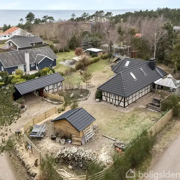 Bindingsværkshus med sort tag står på en grund omkranset af hegn. Ved siden af er flere mindre bygninger og en anlagt have. Kyst udsigt i baggrunden, omgivet af træer.