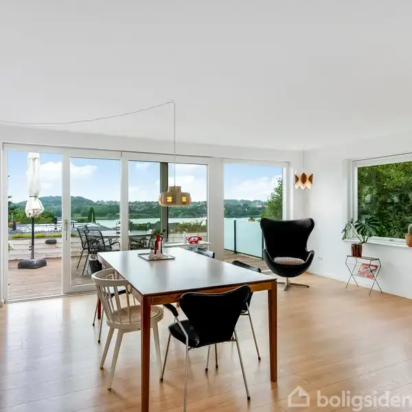 Et spisebord med stole står i et lyst rum med trægulv. I baggrunden er en stor glasdør med udsigt til en terrasse og natur.