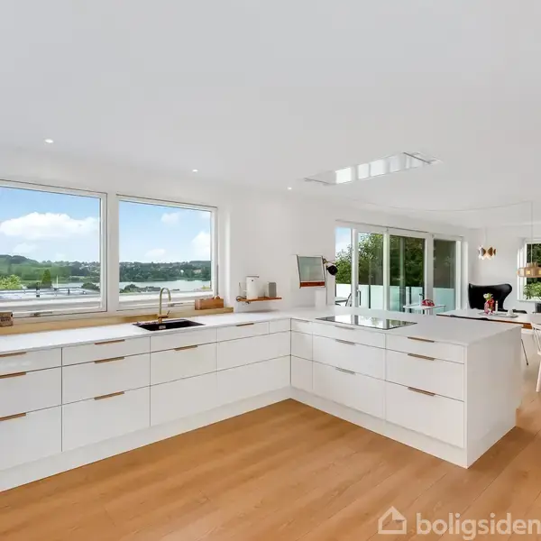 Et moderne køkken med hvide skuffer og ø, der står på et trægulv. Store vinduer med udsigt til natur. Til højre ses en spisestue med borde og stole.