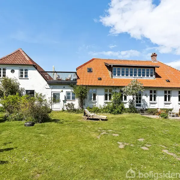 Et hvidt hus med rødt tegltag står i en grøn have med buske og træer. Et trædæk med en liggestol ses foran huset under blå himmel.