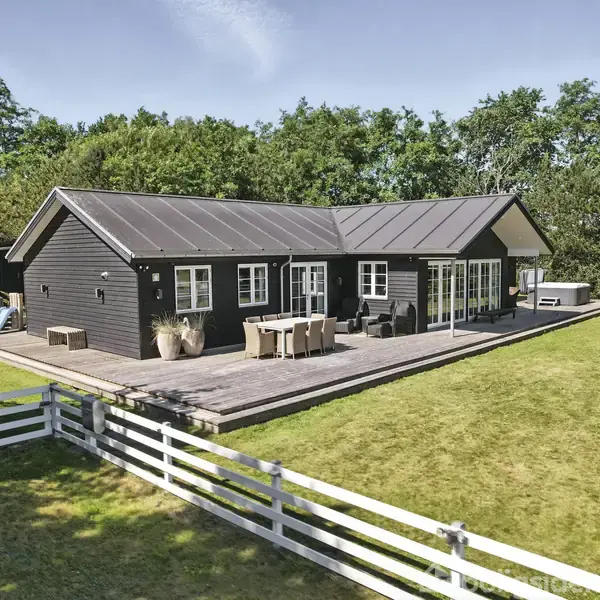 Et sort træhus med terrasse står i en have omgivet af træer. På terrassen er der havemøbler. Græsplæne og hvidt stakit omkranser huset.