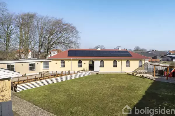 Bygning med solpaneler på taget har en gul facade og store buede vinduer. I forgrunden er en græsplæne omgivet af stier og plankeværk. Træer ses i baggrunden.