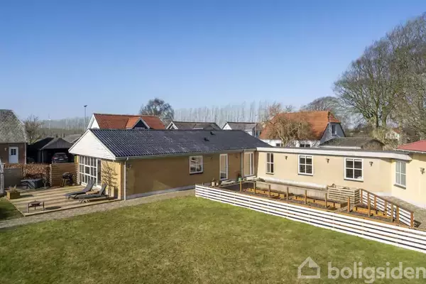 Et enfamiliehus står stille i en velplejet have. Flere terrasser omkranser huset. I baggrunden ses andre huse og træer under en klar blå himmel.