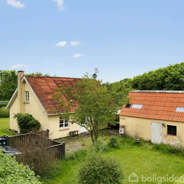 Et gulmalet hus med rødt tag står ved siden af en lade i en frodig have omkranset af buske og træer under en klar blå himmel.