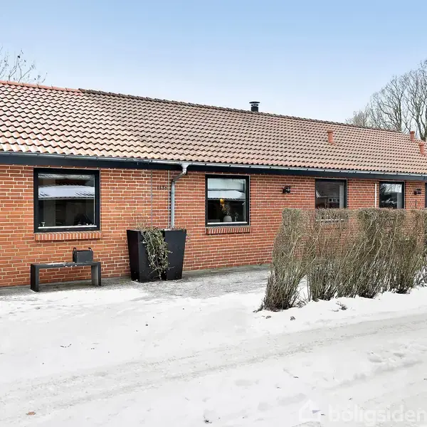En rød murstensvilla med sort tag ligger i et snedækket område. Foran huset er der bar jord og en række buske. Skyfri blå himmel i baggrunden.