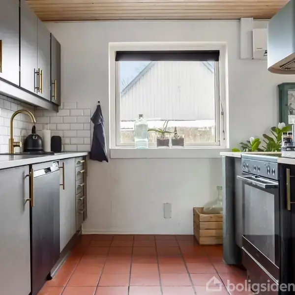 Et moderne køkken med sorte skabe og gyldne håndtag. En elkedel står på bordpladen. Vinduet i midten har udsigt til nabohuset. Planter og en brødkurv dekorerer bordet.