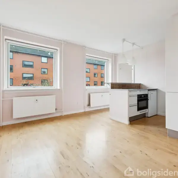 Lyst køkken-alrum med træparketgulv. To store vinduer viser en rød murstensbygning udenfor, mens rummet indeholder et hvidt køkkenmodul med komfur og ovn i en minimalistisk stil.