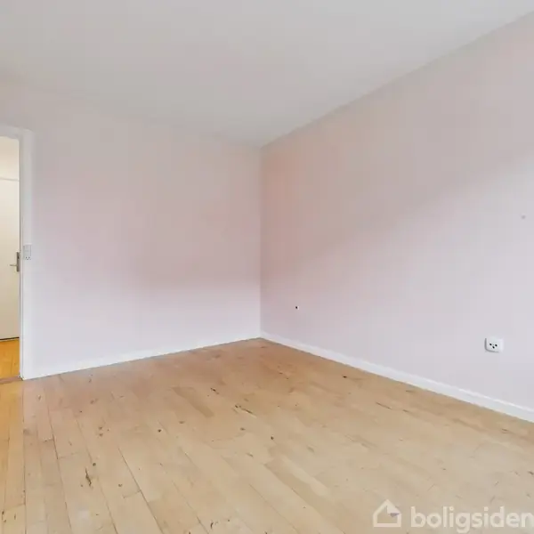 Et tomt værelse med trægulv og lyse vægge. Rummet leder til en lys gang med en dør og postkasse. Sollys strømmer ind fra højre side.