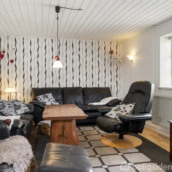 Stue med sorte lædermøbler og et træsofabord, placeret på et mønstret tæppe. Baggrunden er en stribet væg, og rummet oplyses af en loftslampe og et vindue.