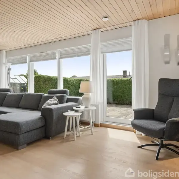 Gråt sofaarrangement står i en lys stue med trægulv og træloft. Store vinduer giver udsigt til en terrasse og en hæk. En høj, grå lænestol står ved siden af lampen.
