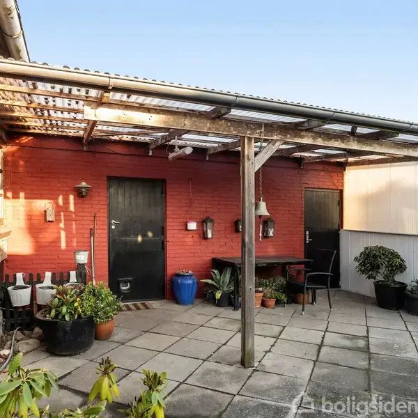 Overdækket terrasse med flisebelægning, adskillige potteplanter i forskellige størrelser langs kanten, rødt murstenshus i baggrunden, og to sorte døre. Sollys skaber skygger på væggen.