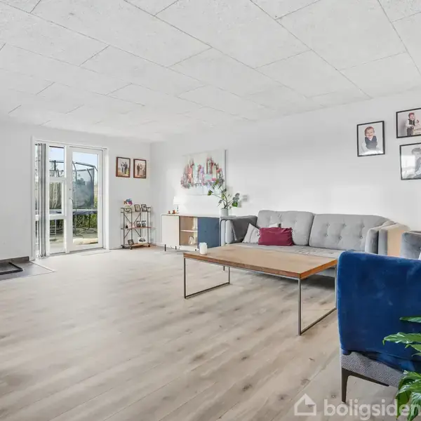 Stue med sofa og sofabord, åbne terrassedøre mod en have. Brændeovn i hjørnet og vægpynt, herunder planter og billeder. Lyse trægulve og minimalistik indretning.