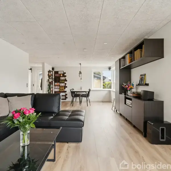 Sort lædersofa står i en lys stue ved siden af et glasbord med blomster. En åben spiseplads med træmøbler ses i baggrunden, moderne indretning omkring.