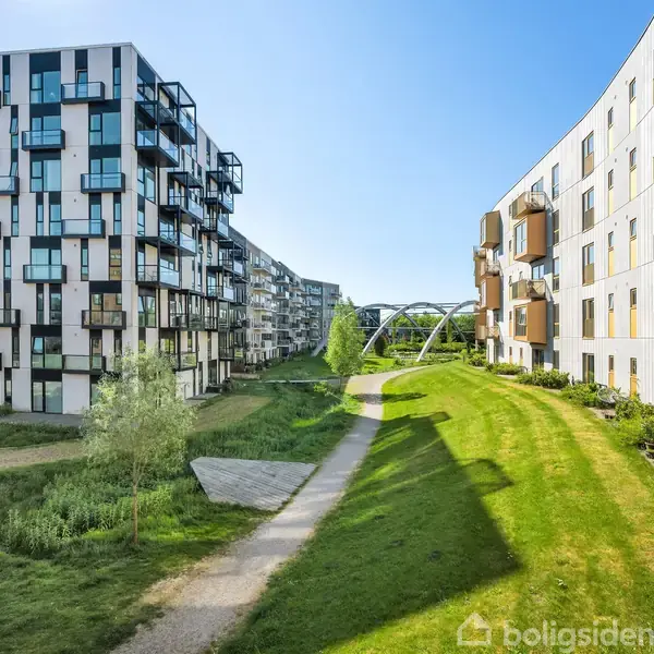 Moderne lejlighedskomplekser står stille langs en snoet sti. I midten en grøn græsplæne og træer, mens en lys blå himmel oplyser den urbane omgivelse.