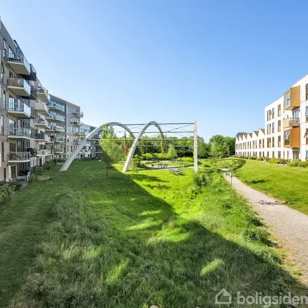 Moderne lejlighedsbygninger står på hver side af en grøn park. I midten passerer en bro med buede søjler. Solen skinner, og en sti løber langs en bygning.