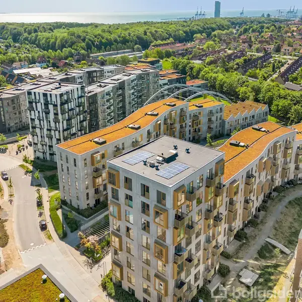Et stort boligkompleks med buede, grønne tage strækker sig gennem en bydel omkranset af skov og bebyggelse, med udsigt til havet i horisonten.
