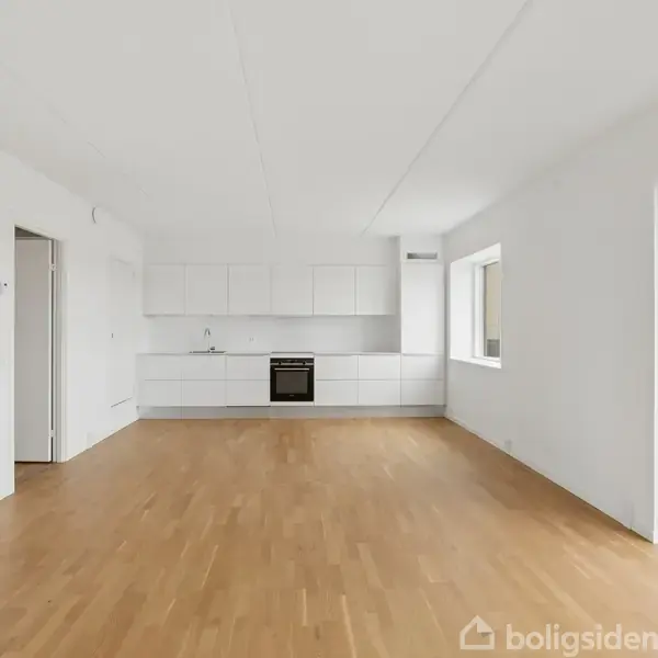 Stue med trægulv og hvidt køkken, tom og rummelig. En åben dør fører til en altan. Vinduer giver naturligt lys, og der er hvide vægge og lofter.