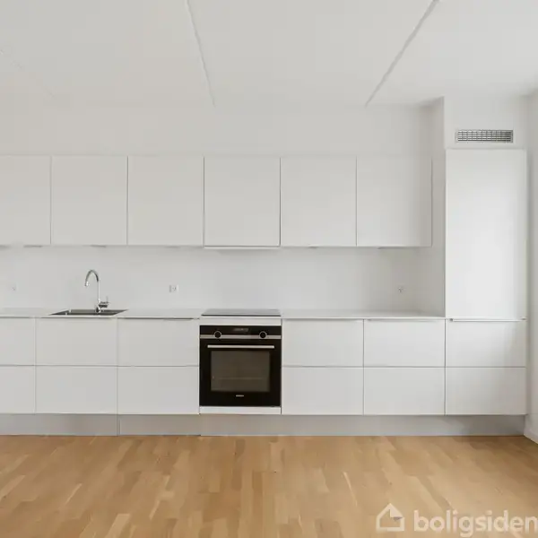 Et moderne køkken med hvide skabe og integreret ovn, set i et lyst værelse med trægulv. Et vindue mod højre viser en udsigt til bygninger udenfor.