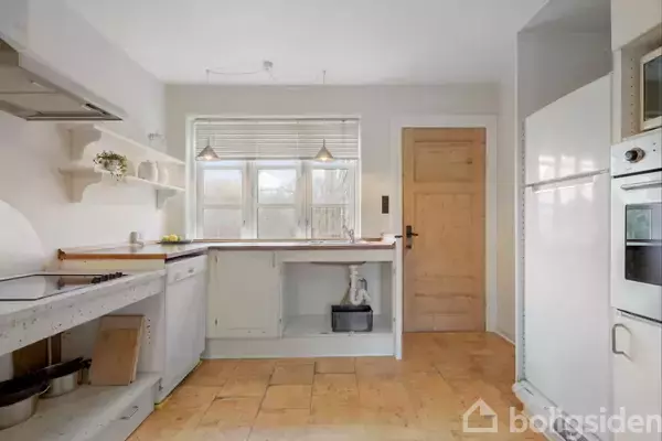 Et lyst køkken med hvide skabe og en høj køleskab-fryser. Trægulv og trædør leder til et andet rum. Vindue over køkkenbordet slipper naturligt lys ind. Hylder med pynt.