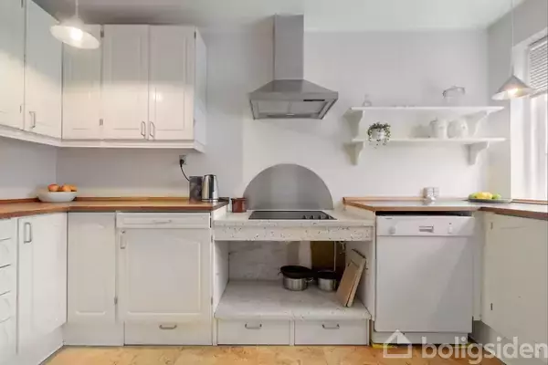 Et lyst køkken med hvide skabe og træbordplader. Komfuret er placeret under en emhætte. Overbord en hylde med vaser og en plante. Køkkenet er ryddeligt og rent.