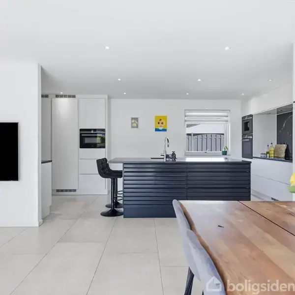 Moderne køkkenområde med sort køkkenø og barstole, står i et lyst rum. Til højre ses et træbord med gul blomsterbuket i en vase. Fladskærms-TV til venstre.