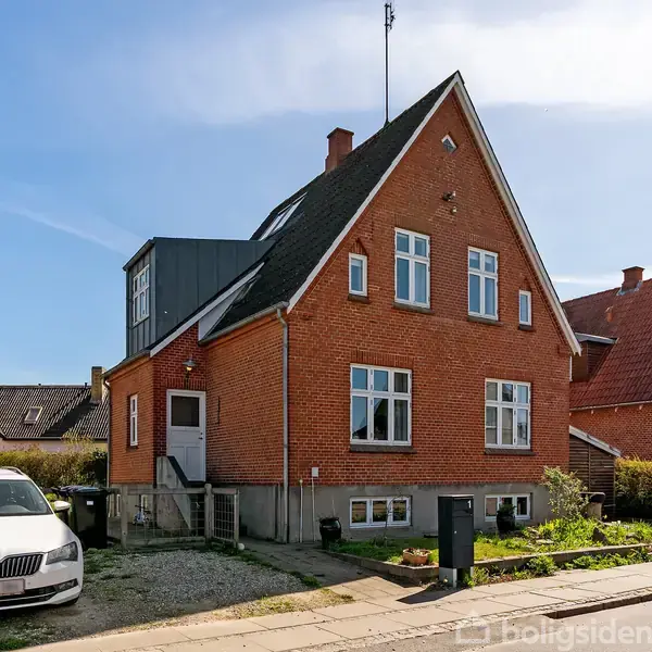 Rødt murstenshus står solid, omgivet af en grøn have og blå himmel. En hvid bil er parkeret til venstre på grusareal ved siden af huset.