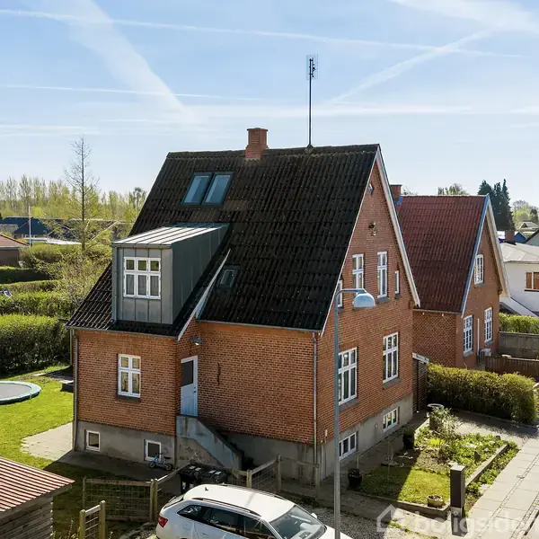 Rødt murstenshus med sort tag har tagvinduer og kvist, placeret i villakvarter med grønne hegn. Trampolin i haven. Parkeret hvid bil ved fortovet. Klar blå himmel.