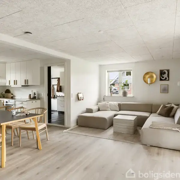 En lys stue med en hjørnesofa og sofabord, der støder op til et køkken med spisebord og stole. Store vinduer slipper naturligt lys ind, omgivet af moderne indretning.