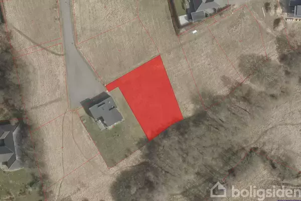 En rød markering viser en grund ved siden af en vej og et hus i et parcelhuskvarter omgivet af træer og åbne marker.