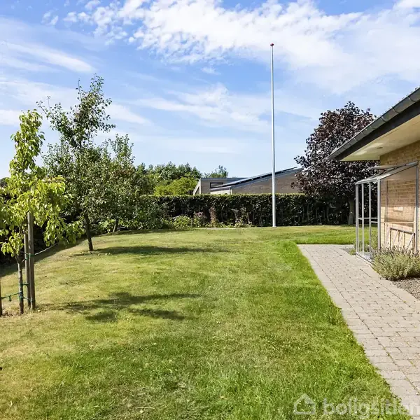 Hus ved siden af en græsplæne med træer i en have. Hækken afgrænser området, og himlen er blå med få skyer.