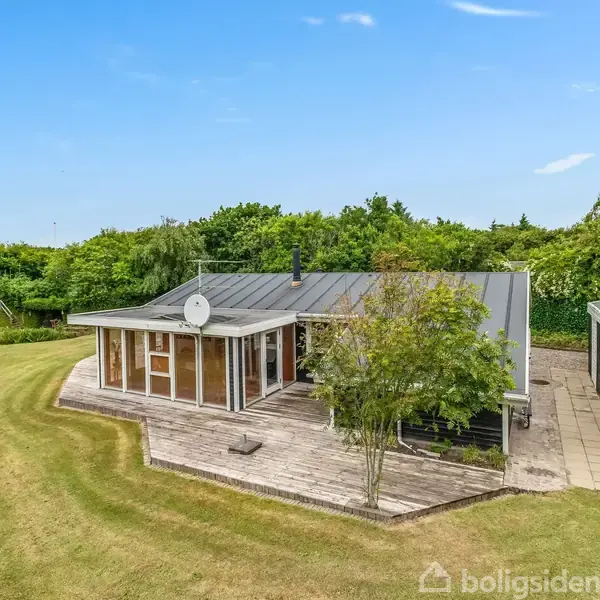 Et sommerhus med store vinduer står på en træterrasse omgivet af en velplejet græsplæne og frodig bevoksning. En carport er til højre for huset.