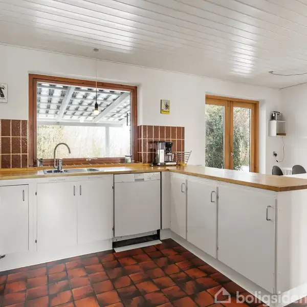 Køkken i hvidt design med træbordplade, kogeplade, opvaskemaskine og vask. Rødt klinkegulv og brun backsplash. Opsat billede og glasdør til spisestue med seks stole ved bordet.