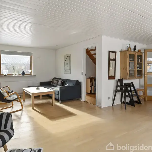 Stue med trægulve, møbleret med en sort sofa, to lænestole og et sofabord. En vindueskarm med dekorationer og en reol ses i baggrunden. En dør leder til et andet rum.