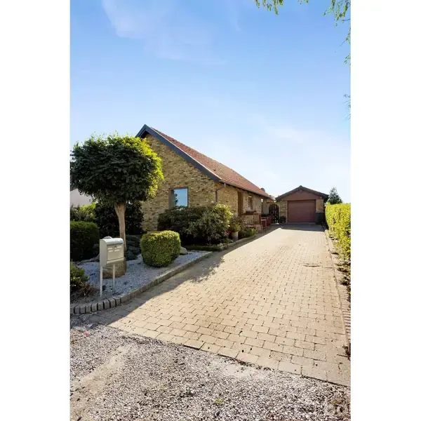 En gulstenshus med rødt tag står langs en flisebelagt indkørsel, omgivet af grønne buske og et træ. En garage ses i baggrunden under en klar blå himmel.