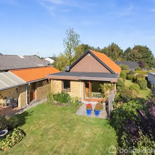 Hus med teglstenstag og store vinduer står i en velholdt have. Grøn græsplæne omgiver huset, mens træer og buske danner en grøn baggrund.