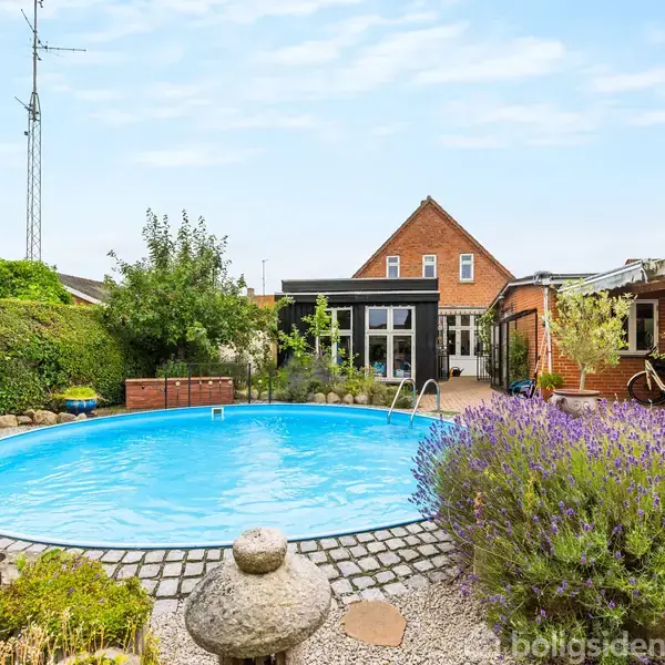 En rund swimmingpool er omgivet af en have med buske og blomster. I baggrunden ses et rødt murstenshus og en overdækket terrasse med grønne planter.