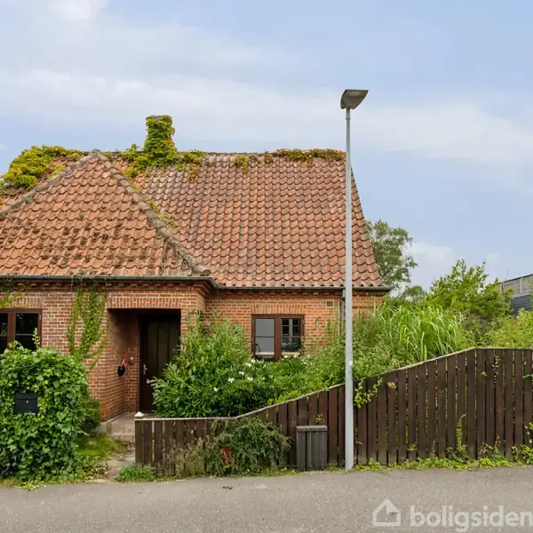 En murstenshus med rødt tegltag dækket af planter, omgivet af en have med frodig bevoksning. En træhegn og en gadelampe står foran huset, omkranset af anden bebyggelse.