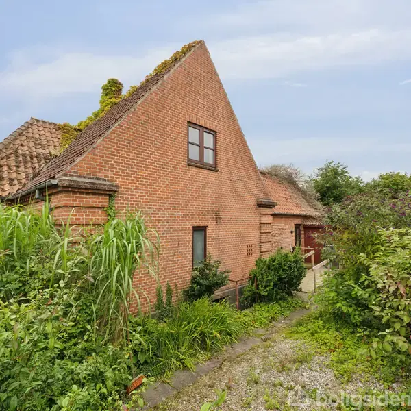 Et rødt murstenshus står omkranset af frodig vegetation. Huset har et spidst tegltag, og vinduerne er placeret asymmetrisk på facaden. Vegetationen skaber en overgroet forhave.