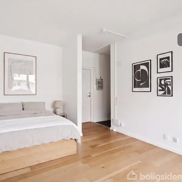 Seng står opredt i et lyst soveværelse med trægulv, flankeret af minimalistiske billeder på væggene, mens en lille lampe står på et natbord ved siden af. Tekst: "Visualisering".