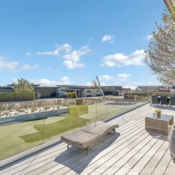 En solrig terrasse med trædæk har solsenge og planter, omgivet af en græsplæne og moderne huse i baggrunden under en himmel med få skyer.