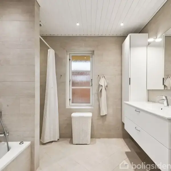 Et moderne badeværelse har et badekar til venstre, en bruseniche med forhæng, og en stor hvid vask med skuffer til højre. Der er et vindue i midten og lys beige fliser.