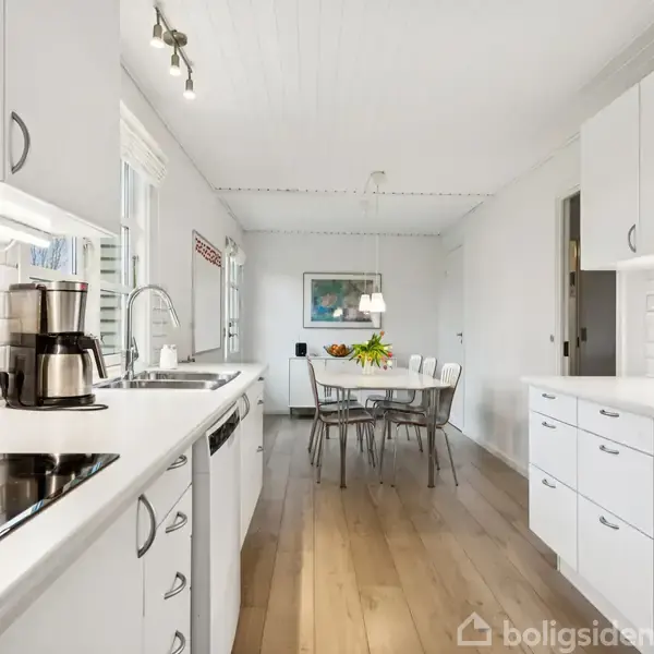 Hvidt køkken med moderne apparater, arrangeret rundt om et bord med stole, mens planter og vægkunst tilfører farve. Trægulve understreger det lyse og luftige rum.