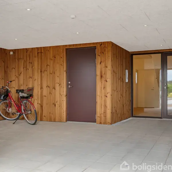 Cykel med kurv står parkeret i en overdækket indgangsparti med træbeklædte vægge. En dør og et stort vindue fører ind til huset. Fliser dækker gulvet.