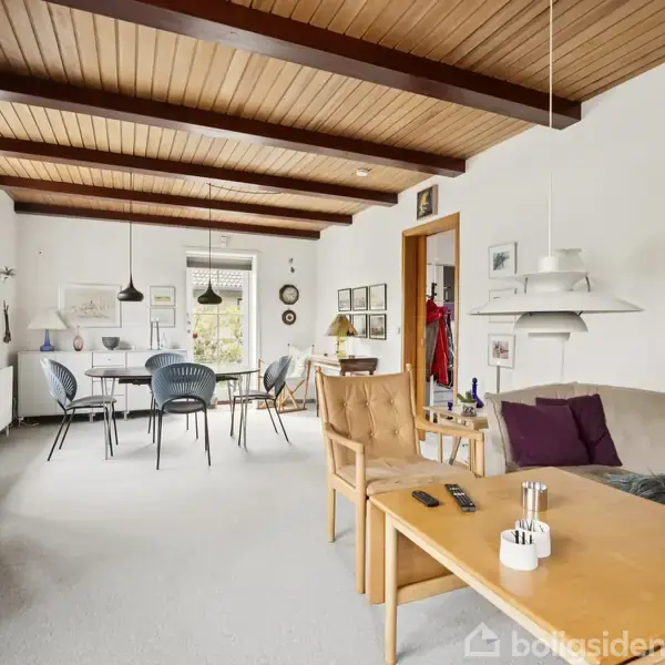 Stue møbleret med bogreol fyldt med bøger langs væggen, spisebord med stole i baggrunden og sofagruppe i forgrunden. Træloft og billeder pryder væggene, skaber en hyggelig atmosfære.