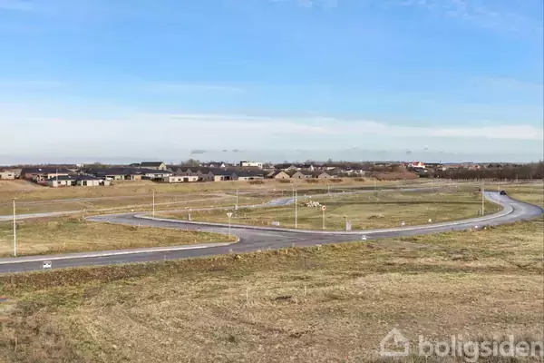 En snoet vej strækker sig gennem et åben landskab af græs med få bygninger i horisonten under en klar blå himmel.