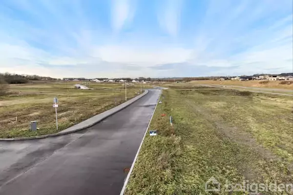 En asfalteret vej løber gennem et fladt, åbent landskab med græs på begge sider, under en blå himmel. Vejskilte og byggegrunde er synlige langs vejkanten.