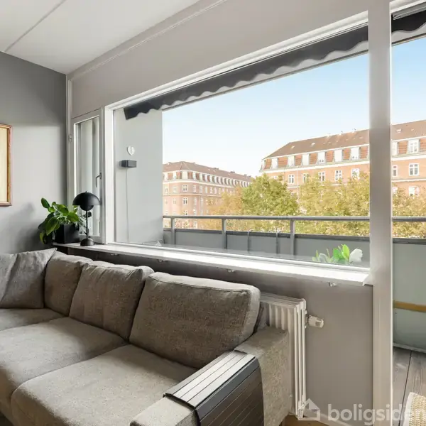 Grå sofa står i stuen ved siden af store vinduer og en åben altandør. Væggen har et gult maleri. Udenfor ses en bygning med mange vinduer.